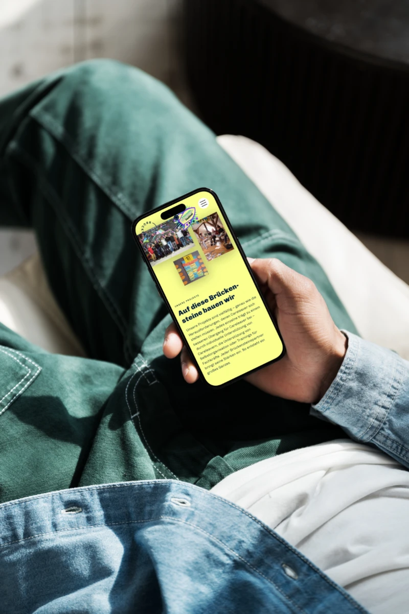 Eine männlich gelesene Person hält ein Smartphone, auf dem Bildschirm ist der Startbildschirm der Website von Brückensteine Careleaver zu sehen.